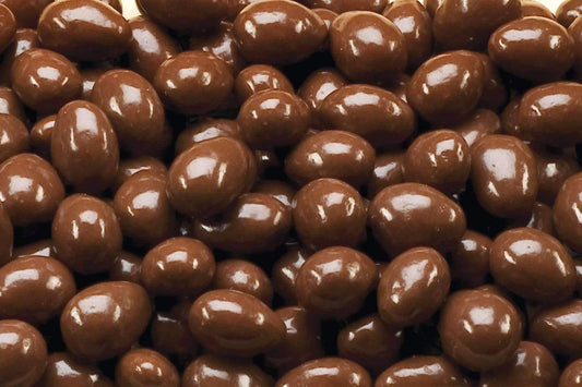 “Close-up of glossy milk chocolate-coated almonds, stacked together on a clean white background showing their smooth texture and rich chocolate coating.”