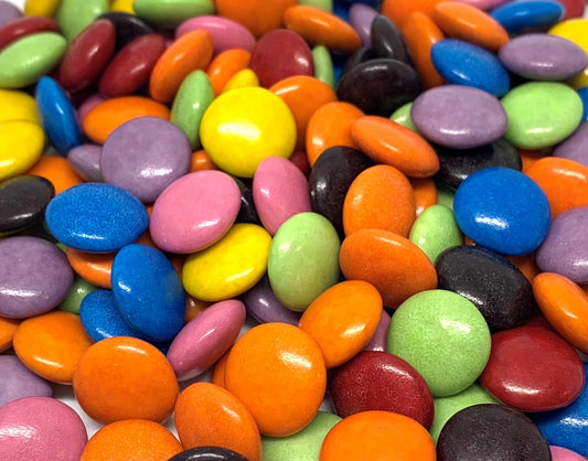 “Close-up of colourful candy-coated chocolate drops in bright rainbow colours, piled together on a clean white background.”
